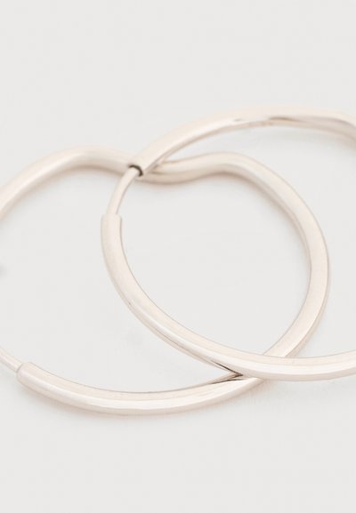 Δύο λαμπερά ασημένια κρίκοι σκουλαρίκια που επικαλύπτονται σε απλό ανοιχτό φόντο.