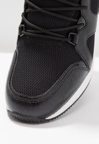 Chaussure de sport noire avec une tige en mesh et des accents noirs brillants. Comprend des détails texturés sur l'avant-pied et une semelle blanche.