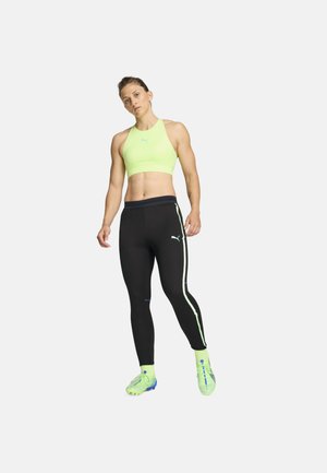 Neon zelena sportska grudnjak s visokim izrezom; crne tajice s bijelim bočnim prugama. Model nosi svijetlo zelene trening tenisice.