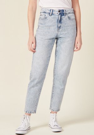 Personne portant un jean taille haute bleu clair et des baskets montantes blanches, debout sur un fond neutre uni.