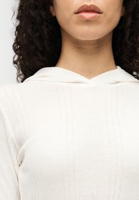 Λευκό πλεκτό πουλόβερ με κουκούλα, με κάθετες ρίγες και υφή. Το ύφασμα φαίνεται απαλό και ελαφρύ.