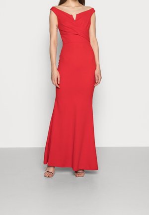 Woman wearing a fitted, off-shoulder red evening gown with a slight sweetheart neckline and floor-length hem, paired with open-toe heels.
