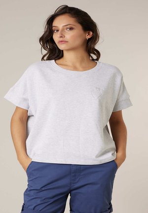 Femme aux cheveux ondulés portant un haut à manches courtes gris clair avec un petit motif en forme de cœur et un pantalon bleu, les mains dans les poches, fond neutre.