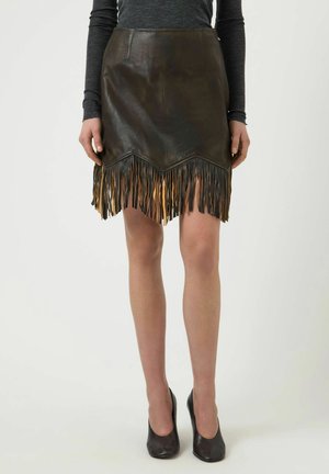 Kvinde iført en mørkebrun lædernederdel med lange frynser og sorte højhælede sko, stående mod en ensfarvet baggrund.