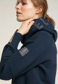 Marinefarbener Hoodie aus strukturiertem Material, mit einer Kapuze mit Kordelzug und einem Aufnäher am Ärmel, auf dem ein Logo zu sehen ist. Streifen-Detail am Kragen.