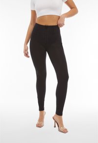 Nainen pukeutuneena korkeavyötäröisiin mustiin leggingsseihin, joissa on edessä vetoketju, ja kirkkaakorkoisiin sandaaleihin, valkoinen lyhyt yläosa, poseeraa käsi lantiolla.