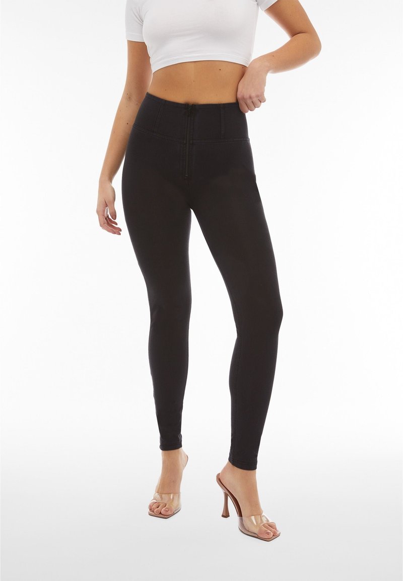 Nainen pukeutuneena korkeavyötäröisiin mustiin leggingsseihin, joissa on edessä vetoketju, ja kirkkaakorkoisiin sandaaleihin, valkoinen lyhyt yläosa, poseeraa käsi lantiolla.