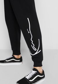 Sort joggebukser med hvid broderet linjekunst på siden. Sammen med sorte sneakers med hvide detaljer og snørebånd.