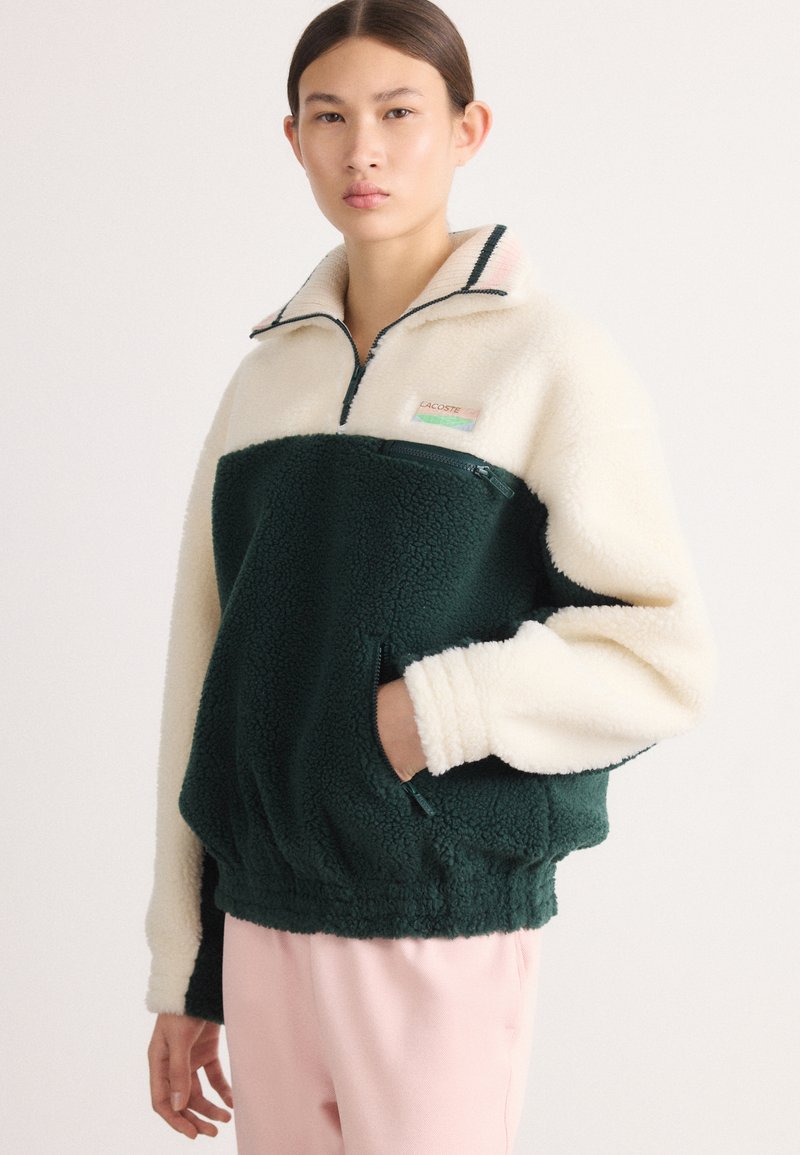 Zeleni i kremasti flis pulover s četvrtim patentnim zatvaračem, koji ima patentni džep, rebrasti ovratnik i malu oznaku s logom na prsima.