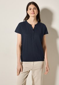 Námořnický modrý top s krátkým rukávem, s knoflíkovým henley výstřihem a uvolněným střihem. Měkký materiál s mírným leskem a detailným nařasením na okraji.