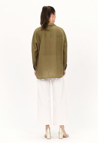 Olivenfarbene, übergroße Bluse mit Kragen, langen Ärmeln und abgerundetem Saum, kombiniert mit weiten weißen Hosen und metallicfarbenen Absatzsandalen.