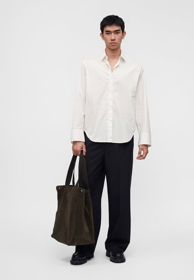 Jeune homme portant une chemise blanche à manches longues et un pantalon noir, tenant un grand sac cabas marron foncé avec des poignées noires, debout sur un fond uni.