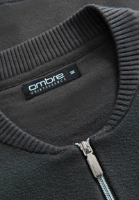 Bindetrøje i mørkegrå stof med ribbet krave. Har en metallisk lynlås og et sort mærke med "ombre QUINTESSENCE" logo.
