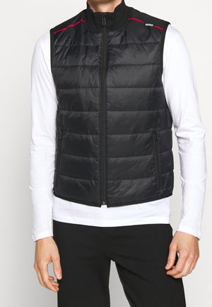 Homme portant une veste matelassée noire zippée sur une chemise blanche à manches longues et un pantalon noir, debout devant un fond uni.