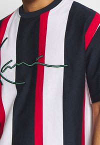 Pánské tričko s krátkým rukávem, pruhované v námořnické modré, červené a bílé barvě z bavlny. Na hrudi má vyšívaný zelený nápis. Standardní kulatý výstřih.