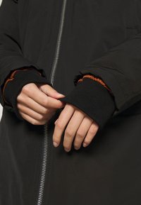 Mains ajustant les poignets d'une veste noire avec des manches intérieures côtelées noires et orange, fermeture éclair argentée visible à l'avant.