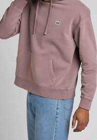 Hooded sweatshirt in mauve, with a kangaroo pocket, ribbed cuffs and hem, featuring a small logo patch on the chest. Made of soft fabric.