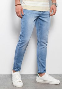 Jeans de mezclilla azul claro con corte Slim, que presentan una textura desvaída, sutiles detalles de costura y puños enrollados sobre zapatillas blancas.