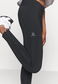 Black athletic leggings with a fitted design, featuring zippered accents, a smooth texture, and a logo on the thigh.