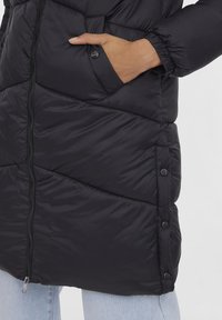 Manteau rembourré noir avec fermeture éclair, coutures diagonales et détails à bouton-pression. Présente une poche frontale et des manches élastiques.
