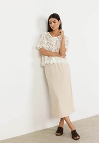 Robe midi en lin beige avec un haut en dentelle crochet blanche, des manches courtes et un ourlet festonné, accompagnée de sandales à glissière marron foncé.