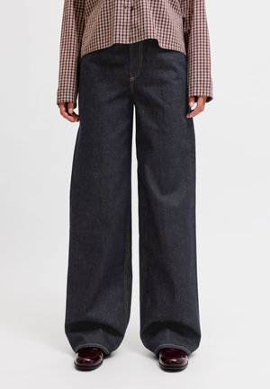 Personne portant un jean large foncé associé à des chaussures marron brillantes et une chemise marron à carreaux à manches longues visible de la taille jusqu'en bas.