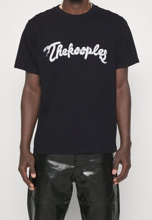 Schwarzes T-Shirt mit kurzen Ärmeln und weißem "The Kooples"-Schriftzug, der Schlangenköpfe zeigt, getragen von einer Person mit glänzenden schwarzen Hosen vor einfarbigem Hintergrund.