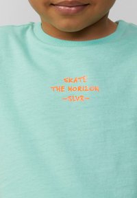 Helles Türkis Sweatshirt mit geripptem Rundhalsausschnitt. Mit orangefarbenem, gesticktem Text: "SKATE THE HORIZON -SLVR-." Weicher, glatter Stoff.