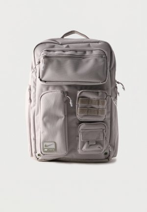 Grauer rechteckiger Rucksack mit mehreren vorderen Reißverschlusstaschen und einem oberen Tragegriff, mit einem kleinen Nike Utility 2.0 Logo-Patch.