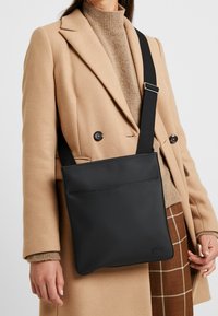 Personne portant un manteau beige et une jupe à carreaux porte un sac bandoulière noir avec une sangle réglable sur l'épaule.
