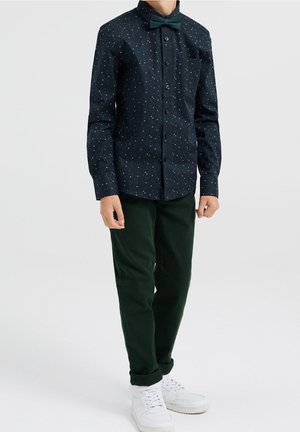 Donkerblauwe overhemd met kleine kleurrijke patronen, gecombineerd met donkergroene broek en witte sneakers. Strikje voegt een formele toets toe.