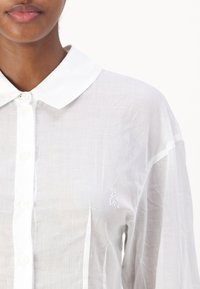 Chemise blanche à manches longues avec un col, présentant une texture légère et un subtil accent brodé sur la gauche ; manches longues avec fentes sur les côtés.