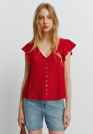Ung kvinde med langt lyst hår iført en rød bluse med knapper og flæseærmer samt lyseblå denimshorts, der holder et brunt håndtag.