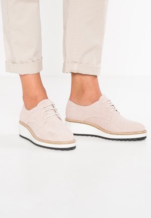 Lyse beige teksturerede snøresko med plateau, båret med opkrøllede lyse beige bukser på en hvid gulvbaggrund.