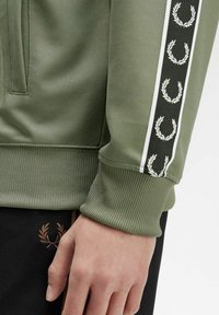 Giacca sportiva verde con polsini a costine, presenta una banda nera con motivi di alloro bianchi. Il materiale ha una finitura testurizzata.