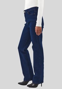 Osoba oblečená v tmavě modrých rovně střižených džínách, bílém topu s dlouhým rukávem a černých špičatých lodičkách na vysokém podpatku s kovovou sponou.