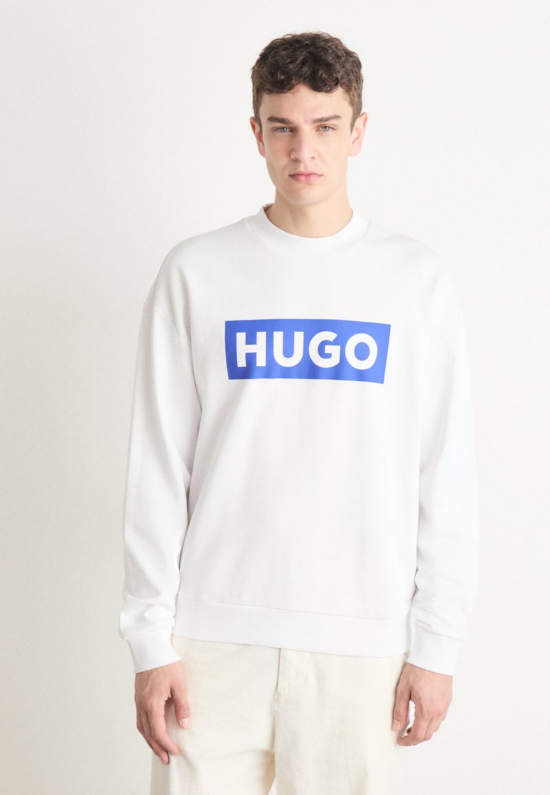 Balta ilgomis rankovėmis džemperis su mėlynu stačiakampiu logotipu, kuriame užrašyta "HUGO" balta spalva. Minkšta tekstūra, raukšlių apykaklė ir rankogaliai.