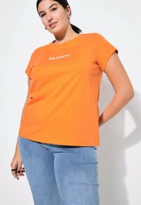 Orange Baumwoll-T-Shirt mit kurzen Ärmeln, auf dem "MÜNCHEN" in Weiß über der Brust gedruckt ist. Kombiniert mit hellblauen Jeans.