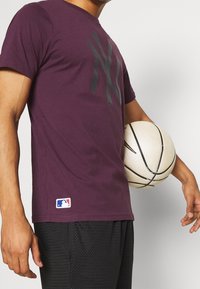 Mörkröd bomullströja med en svart logotyp på framsidan, kortärmad, och MLB-märke på nederkanten; bärs med svarta meshshorts och håller i en basketboll.