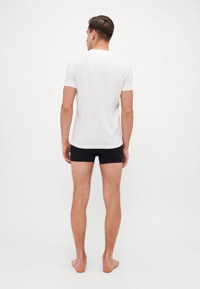 Weißes T-Shirt mit kurzen Ärmeln und schwarze Boxershorts, getragen von einem männlichen Model, von hinten gesehen, barfuß vor einem schlichten Hintergrund.