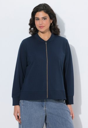 Frau mit lockigen dunklen Haaren, die goldene Creolen, eine marineblaue Jacke mit Reißverschluss und hellblaue Jeans trägt, vor einem weißen Hintergrund stehend.