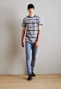 Siva prugasta majica s kratkim rukavima, svijetloplave uske hlače i crne cipele. Model nosi crnu kapu i stoji pred jednostavnom pozadinom.