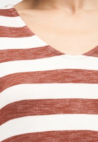 Κοντινό πλάνο στο πάνω μέρος του στήθους και του λαιμού ενός ατόμου που φοράει καφέ και λευκή ριγέ μπλούζα με V λαιμόκοψη.