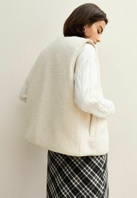 Krämfärgad fleeceväst med en boxig design, utan knappar, och sidofickor, bärs över en vit kabelstickad tröja och rutiga kjol.