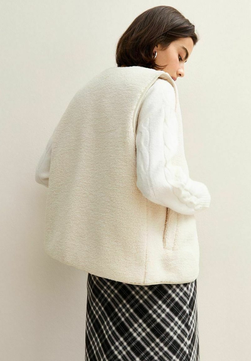 Krämfärgad fleeceväst med en boxig design, utan knappar, och sidofickor, bärs över en vit kabelstickad tröja och rutiga kjol.