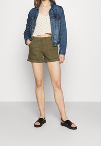 Femme portant une veste en denim bleue, un crop top en tricot crème, un short vert olive et des sandales noires, debout devant un fond blanc uni.