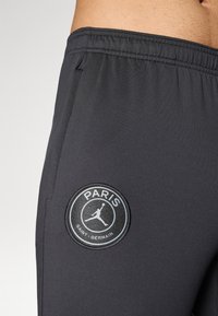 Sorteidene treningsbukser i svart med en glatt tekstur, elastisk midje og en rund logo-patch med "PARIS" og "SAINT-GERMAIN" i hvitt.