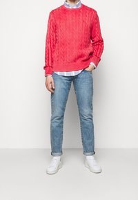 Persona in piedi con la mano in tasca, che indossa un maglione corallo a treccia sopra una camicia a righe blu, jeans azzurri chiari, sneakers bianche e calzini.