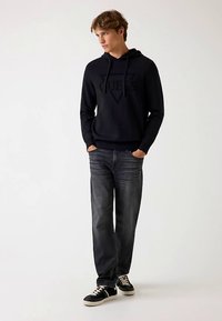 Must kapuutsiga pusa, millel on reljeefne logo, ühendatud tumedate teksade ja mustade tossudega. Modell poseerib käed taskutes heleda tausta ees.