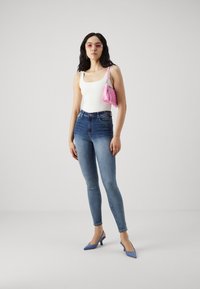 Jeans azules ajustados, top blanco sin mangas, bolso rosa, tacones azules. Lleva gafas de sol transparentes rosadas, cabello oscuro ondulado, accesorios mínimos.